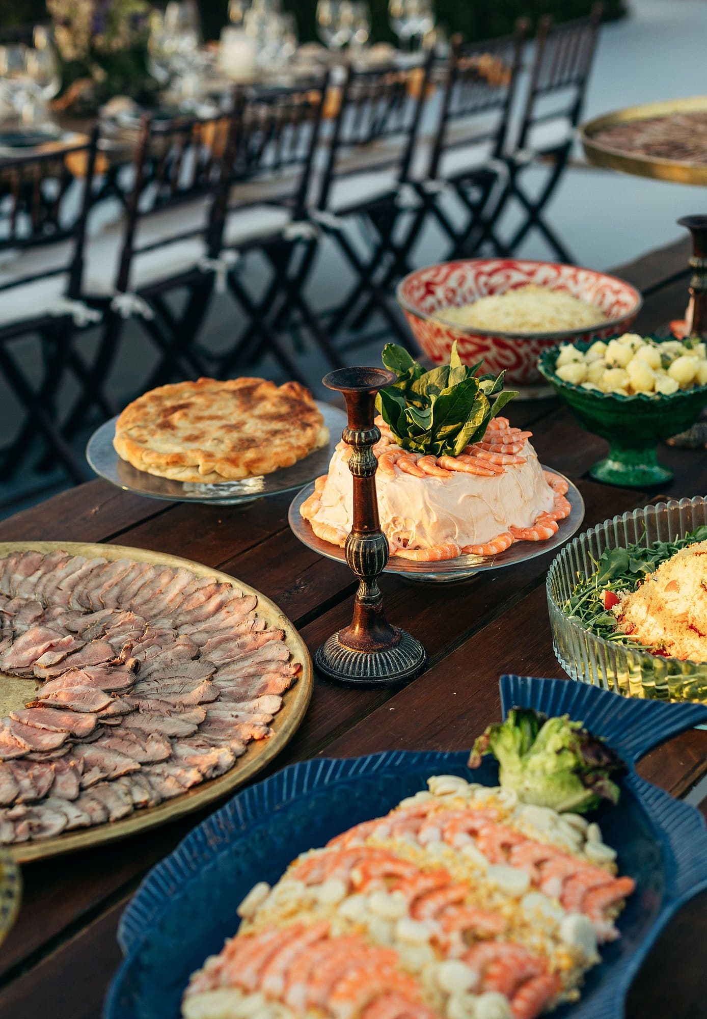 Travessas de carnes frias, marisco e salgados, com a mesa dos convidados posta ao fundo.