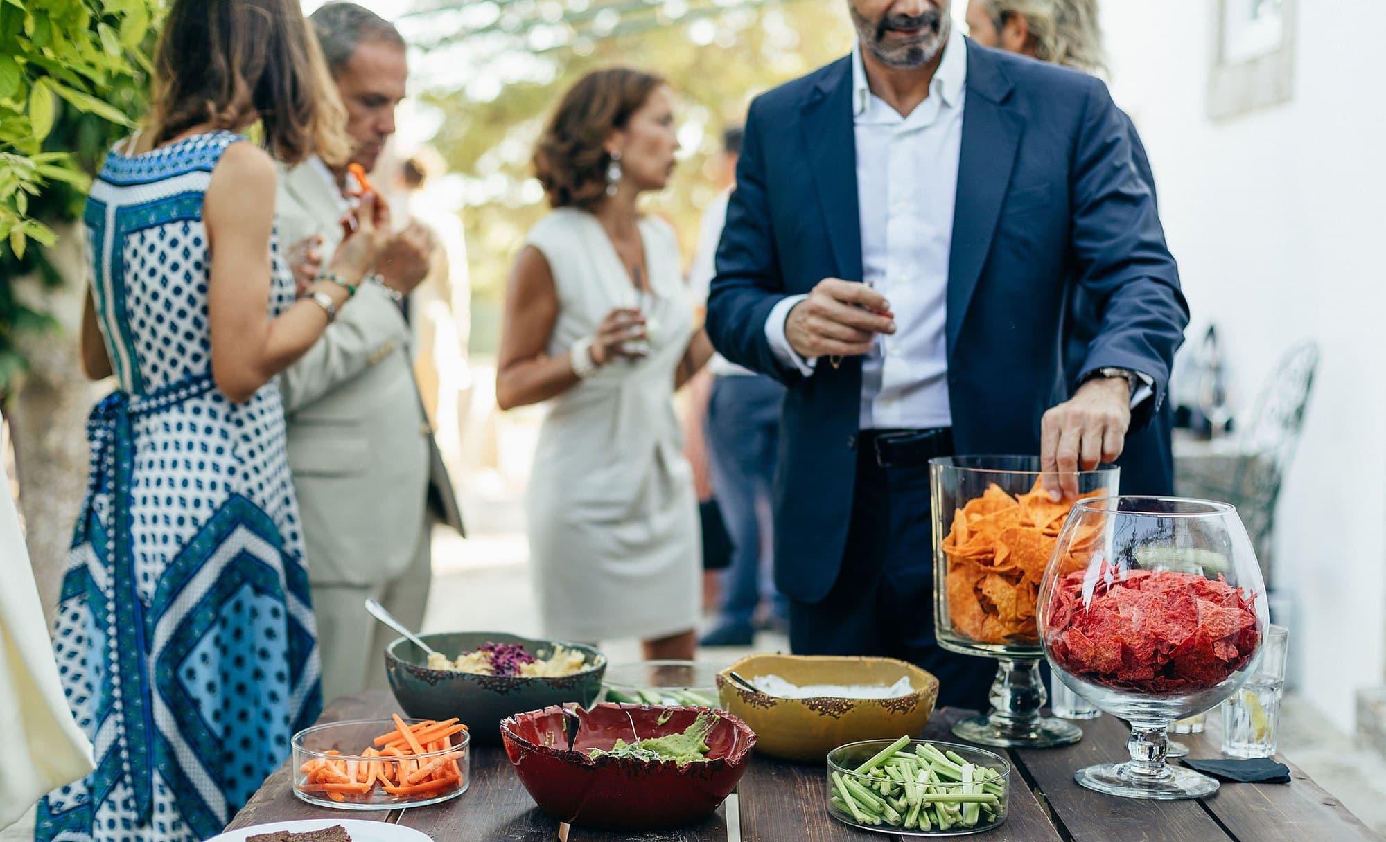 Convidados à volta de uma estação de petiscos, com crudités, pastas e chips de legumes.